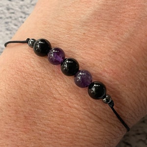 Puede incluir: Un brazalete de cordón negro con cuentas negras y moradas alternas. El brazalete está atado con un nudo.