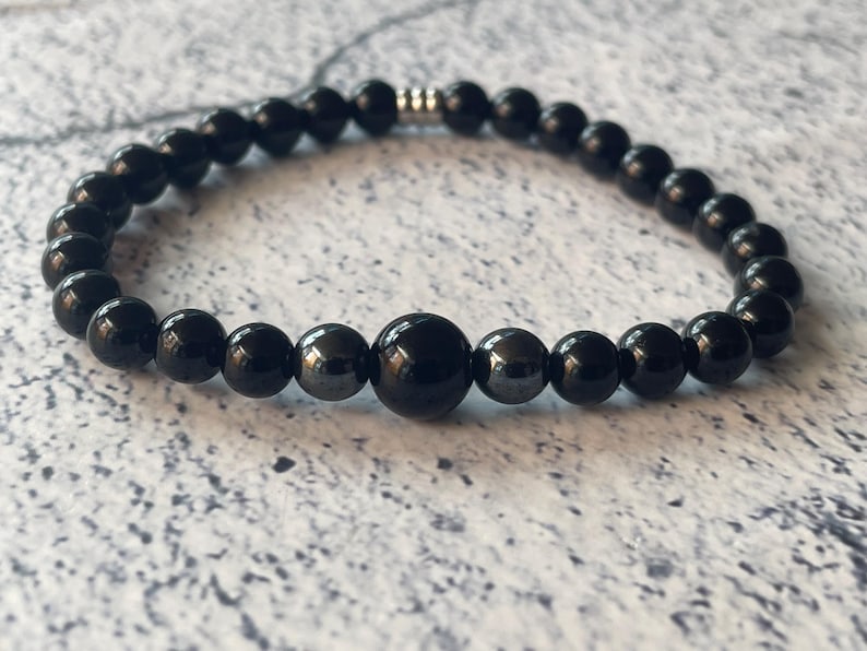 May include: Black beaded bracelet with a silver clasp. The bracelet is made of round black beads and has a single larger black bead in the center.