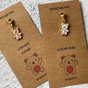 May include: Two brown cards with gold charms attached. The charms are of cartoon bears with red hearts. The cards have the text "Sending you" and "Love and a hug!" on one card and "Sending you" and "A bear hug!" on the other card. The cards are signed "By Christi."
