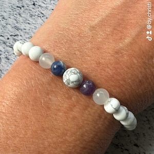May include: A beaded bracelet featuring a sequence of white marble-patterned beads, translucent clear beads, a single dark blue bead, and a single purple bead. The beads are strung together in a circular pattern to form a flexible wrist accessory.