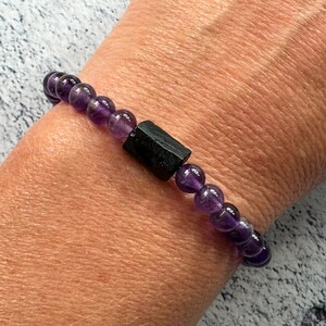 May include: A purple amethyst gemstone bracelet with a black obsidian bead. The bracelet is made with round beads and is worn on a person's wrist.
