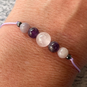May include: A purple and white beaded bracelet with a lavender cord. The bracelet features amethyst, rose quartz, and moonstone beads.