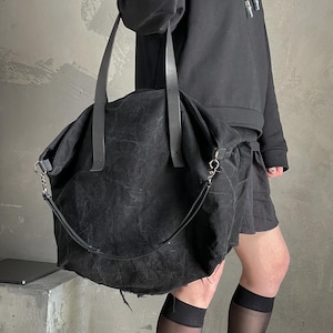 May include: A large, slouchy black tote bag crafted from a textured, distressed fabric. The bag features two long, flat shoulder straps and a detachable, thinner crossbody strap attached with silver-toned metal hardware clips on both sides.