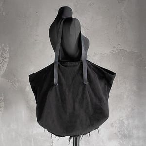 May include: A black tote bag with long straps, displayed on a black mannequin. The bag has a rounded bottom and a frayed edge. The background is a textured gray wall.