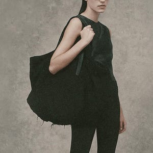 May include: A person wearing black sunglasses, a black sleeveless top, and flared black pants. They are carrying a large black tote bag over their shoulder. The background is a solid, neutral gray.