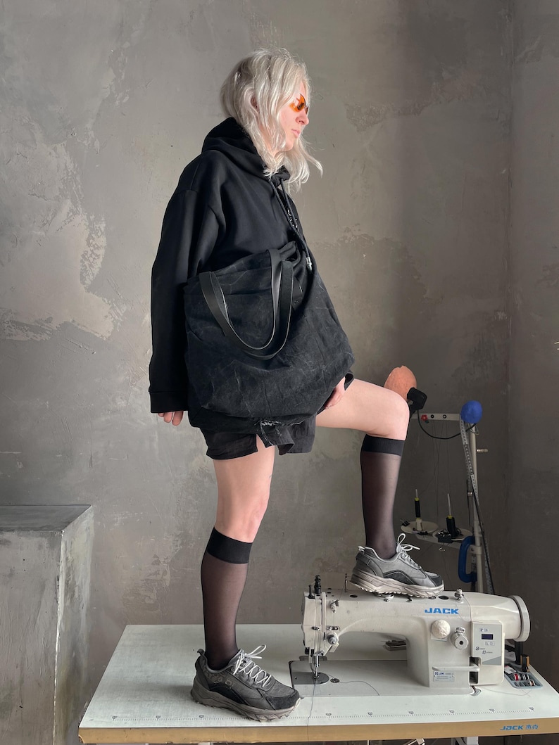 May include: A black hooded sweatshirt, black shorts, and a large black tote bag. The outfit includes sheer black knee-high socks and grey athletic trainers. The person is standing with one foot on a grey industrial sewing machine labelled JACK.