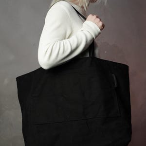 May include: A large black tote bag with a rectangular shape and a front pocket. The bag is carried over the shoulder by a person wearing a white waffle-knit long-sleeve shirt.