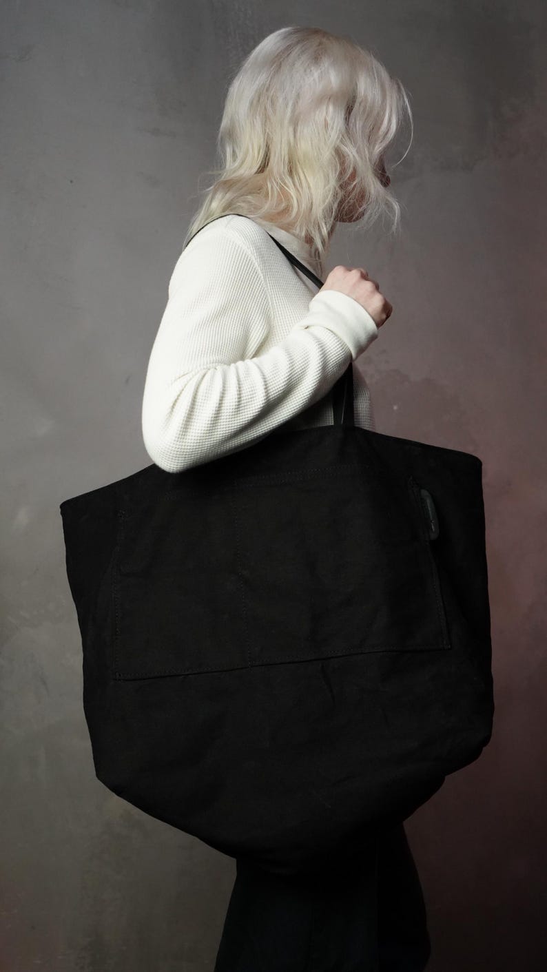 May include: A large black tote bag with a rectangular shape and a front pocket. The bag is carried over the shoulder by a person wearing a white waffle-knit long-sleeve shirt.