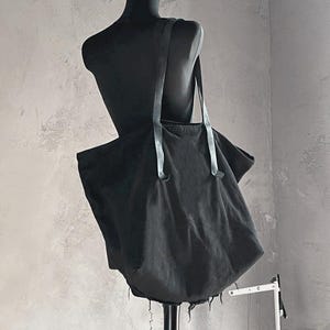 May include: A black tote bag with long straps is displayed on a black mannequin. The bag is made of a soft, dark fabric and has a raw, frayed edge along the bottom. The straps are a lighter shade of black.