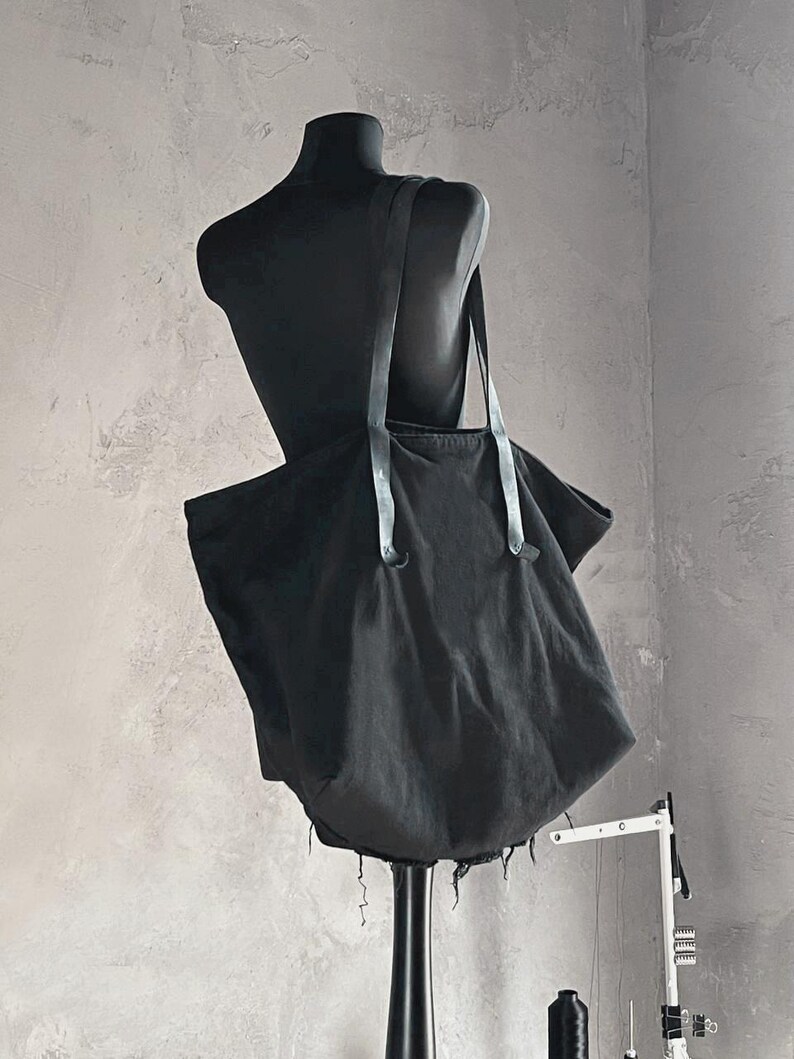 May include: A black tote bag with long straps is displayed on a black mannequin. The bag is made of a soft, dark fabric and has a raw, frayed edge along the bottom. The straps are a lighter shade of black.