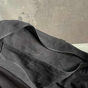 May include: A black canvas bag featuring a large main compartment with a secure zip closure. The bag includes an exterior slip pocket along the side, constructed from the same black fabric, providing additional storage space for small items.