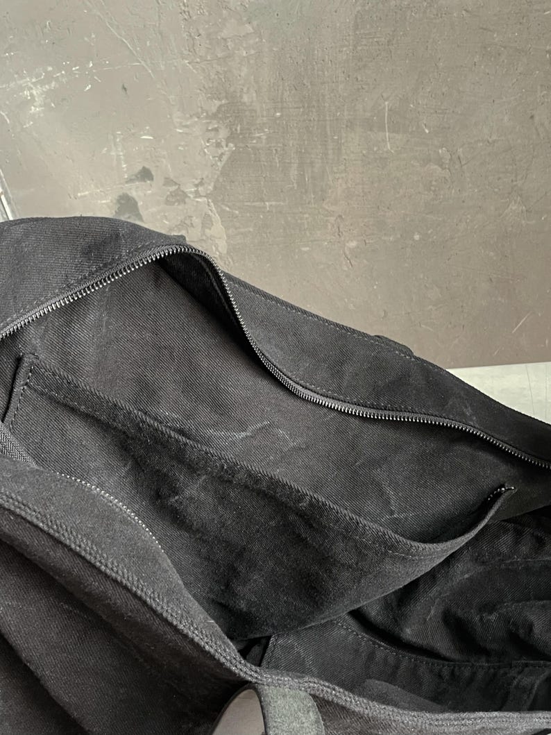 May include: A black canvas bag featuring a large main compartment with a secure zip closure. The bag includes an exterior slip pocket along the side, constructed from the same black fabric, providing additional storage space for small items.