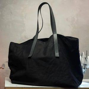 May include: A large black tote bag with long, dark leather handles. The bag is made of a soft, textured fabric and appears to be empty. The handles are attached with small, dark leather accents. The bag is sitting on a white surface.