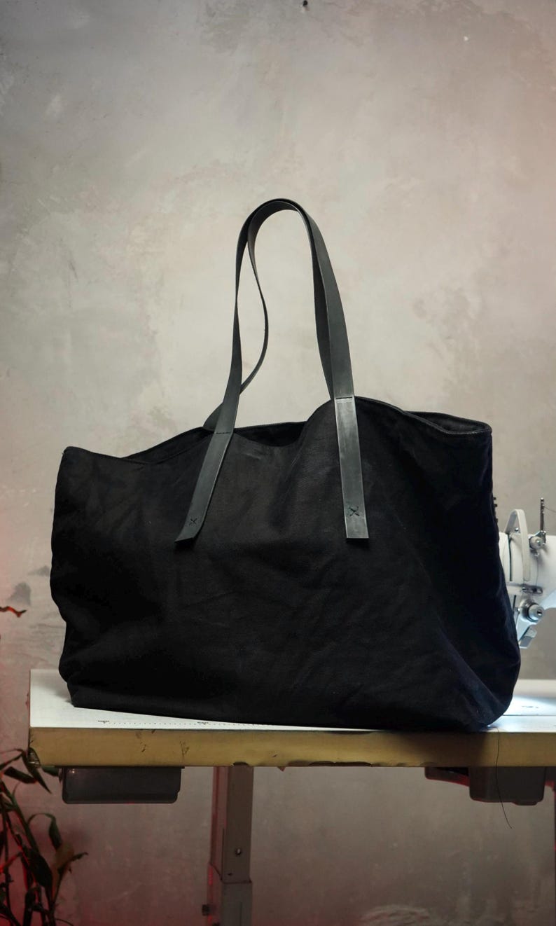 May include: A large black tote bag with long, dark leather handles. The bag is made of a soft, textured fabric and appears to be empty. The handles are attached with small, dark leather accents. The bag is sitting on a white surface.