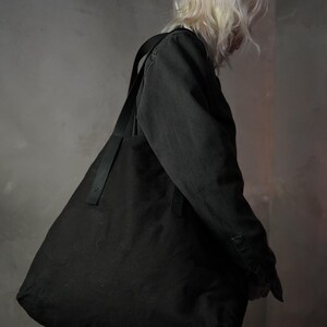 May include: A large, black tote bag with a long, black strap is worn over the shoulder. The bag is made of a soft material and has a frayed edge. The person wearing the bag is facing away from the camera.