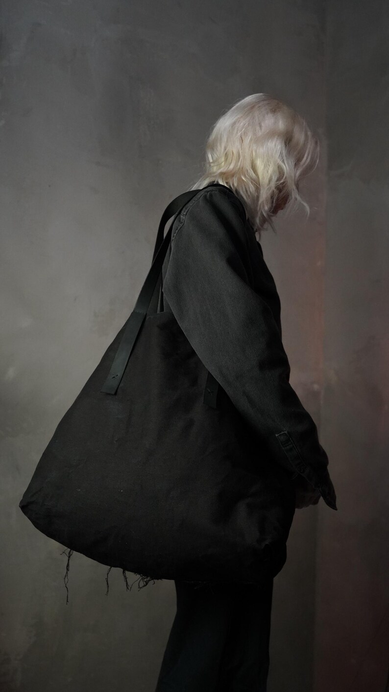 May include: A large, black tote bag with a long, black strap is worn over the shoulder. The bag is made of a soft material and has a frayed edge. The person wearing the bag is facing away from the camera.