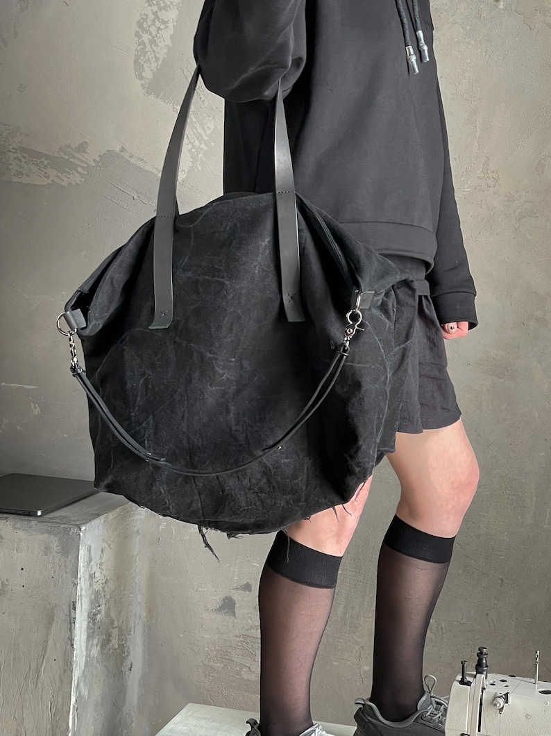 May include: A large, slouchy black tote bag crafted from a textured, distressed fabric. The bag features two long, flat shoulder straps and a detachable, thinner crossbody strap attached with silver-toned metal hardware clips on both sides.