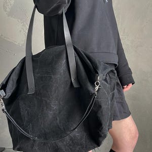 May include: A large black tote bag crafted from a textured, distressed fabric. The bag features two long, flat shoulder straps and a detachable, thinner crossbody strap with a silver-toned metal lobster clasp attachment on the side.