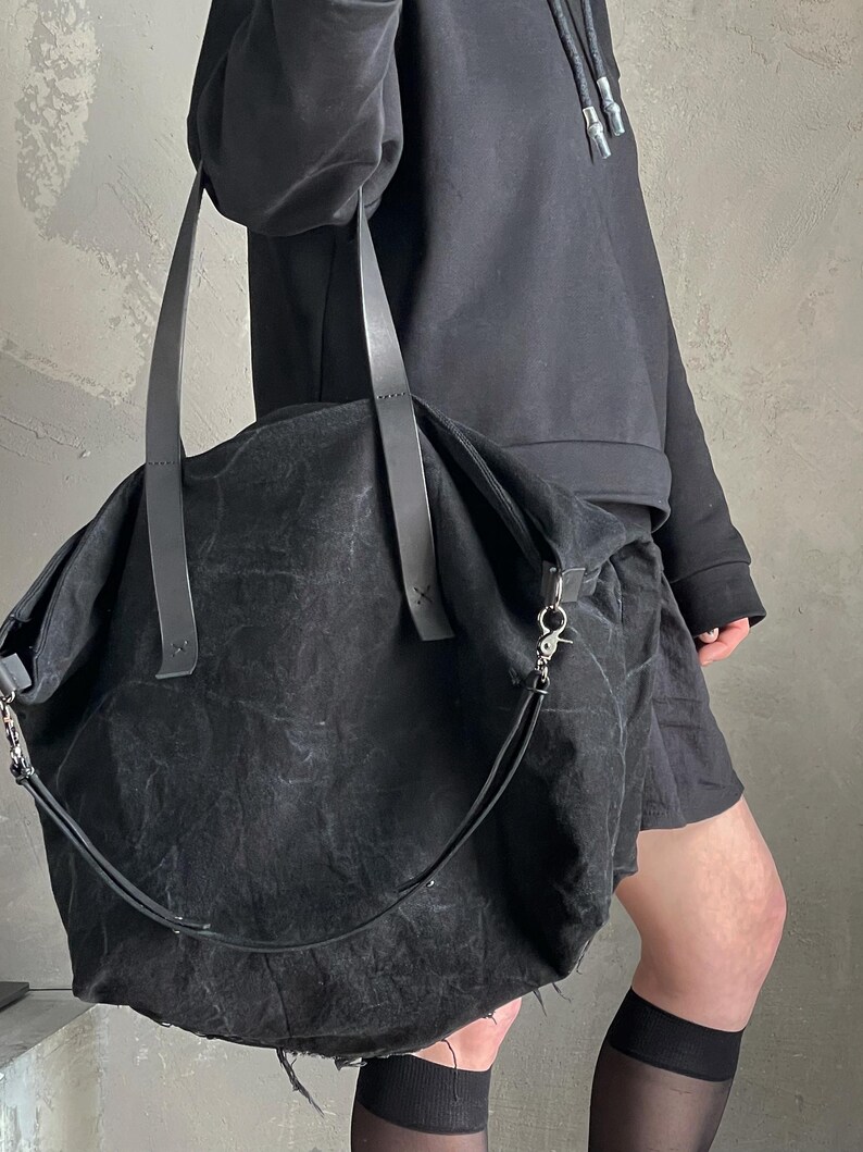 May include: A large black tote bag crafted from a textured, distressed fabric. The bag features two long, flat shoulder straps and a detachable, thinner crossbody strap with a silver-toned metal lobster clasp attachment on the side.