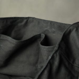 May include: Close-up of a dark gray tote bag with multiple pockets. The bag is made of a sturdy fabric, with visible stitching along the edges. The interior pockets are visible, suggesting ample storage space. The bag's design is simple and functional.