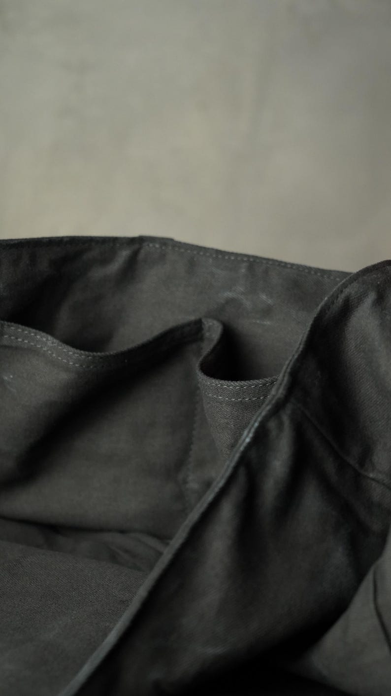 May include: Close-up of a dark gray tote bag with multiple pockets. The bag is made of a sturdy fabric, with visible stitching along the edges. The interior pockets are visible, suggesting ample storage space. The bag's design is simple and functional.