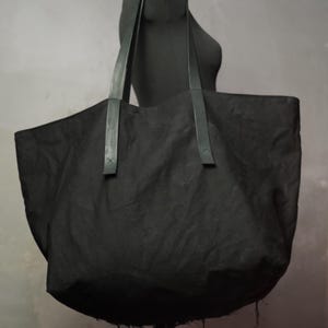 May include: A large, black tote bag with long, dark handles. The bag is made of a textured fabric and is displayed on a black mannequin. The bag has a wide opening and a rounded bottom, suitable for carrying various items.