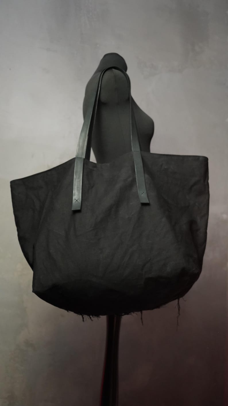 May include: A large, black tote bag with long, dark handles. The bag is made of a textured fabric and is displayed on a black mannequin. The bag has a wide opening and a rounded bottom, suitable for carrying various items.