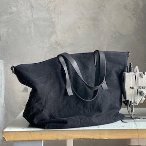 May include: A black canvas tote bag featuring two dark leather shoulder straps attached with visible stitching. The bag has a wide, rectangular body with a soft, unstructured shape and is positioned next to a sewing machine on a work table.