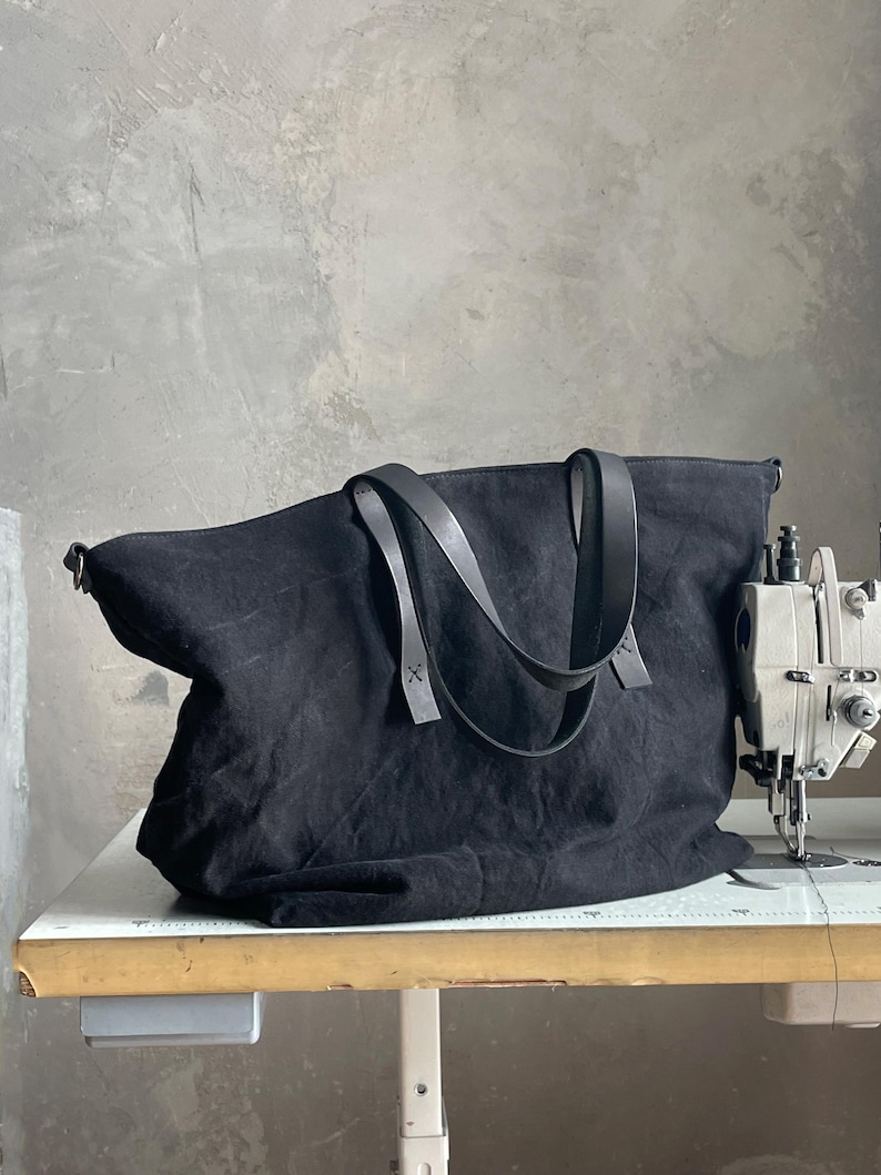 May include: A black canvas tote bag featuring two dark leather shoulder straps attached with visible stitching. The bag has a wide, rectangular body with a soft, unstructured shape and is positioned next to a sewing machine on a work table.
