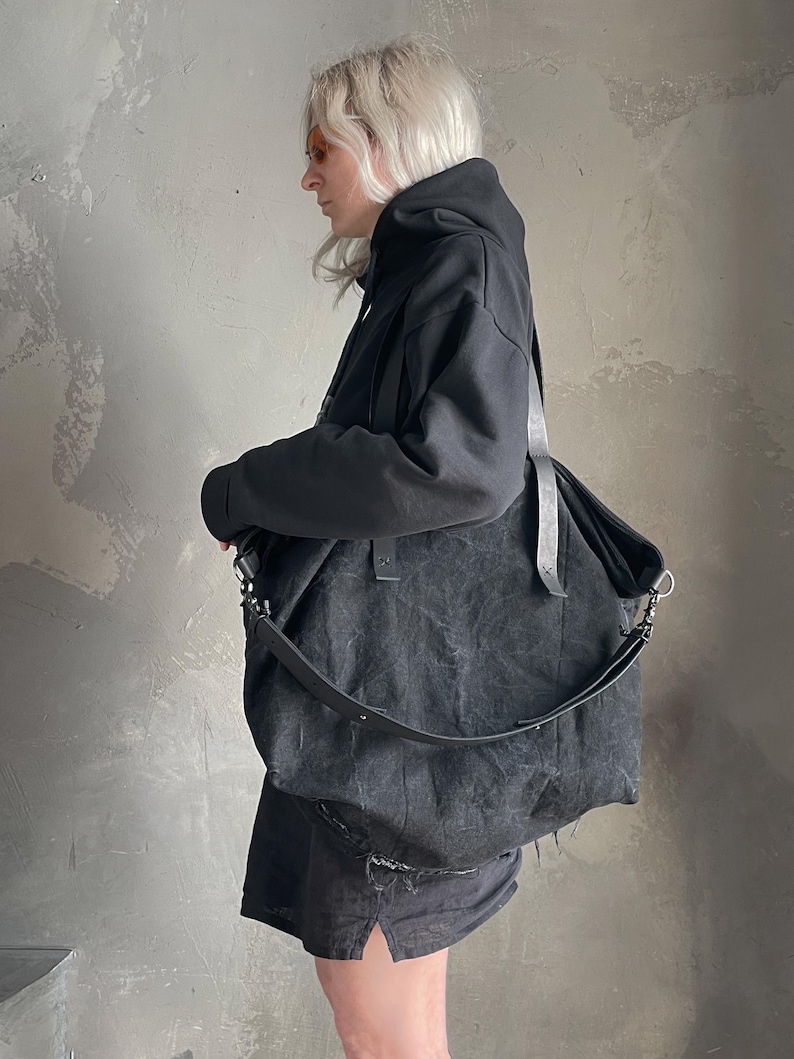 May include: A large, slouchy black shoulder bag with a textured, distressed finish. The bag features two long black straps, silver-toned metal hardware, and a wide, adjustable crossbody strap attached with metal clips on the sides.