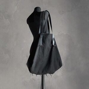 May include: A black tote bag with leather straps is displayed on a black mannequin. The bag has a simple design with a raw, frayed edge at the bottom. The background is a textured gray wall.