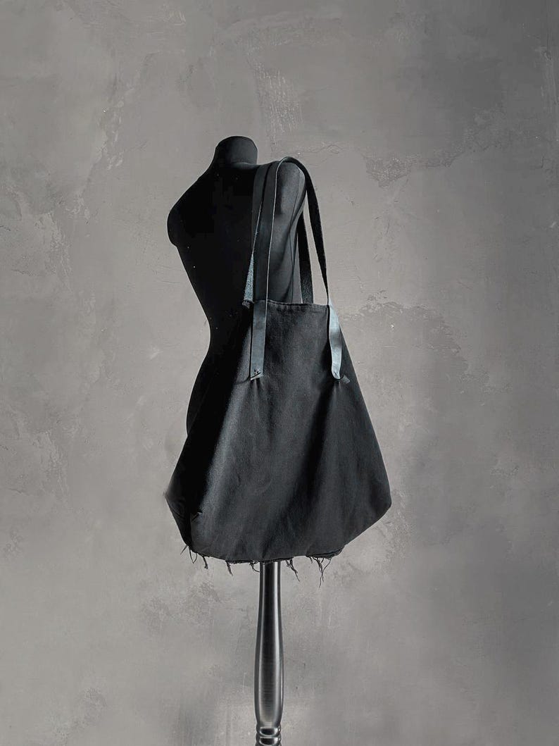 May include: A black tote bag with leather straps is displayed on a black mannequin. The bag has a simple design with a raw, frayed edge at the bottom. The background is a textured gray wall.