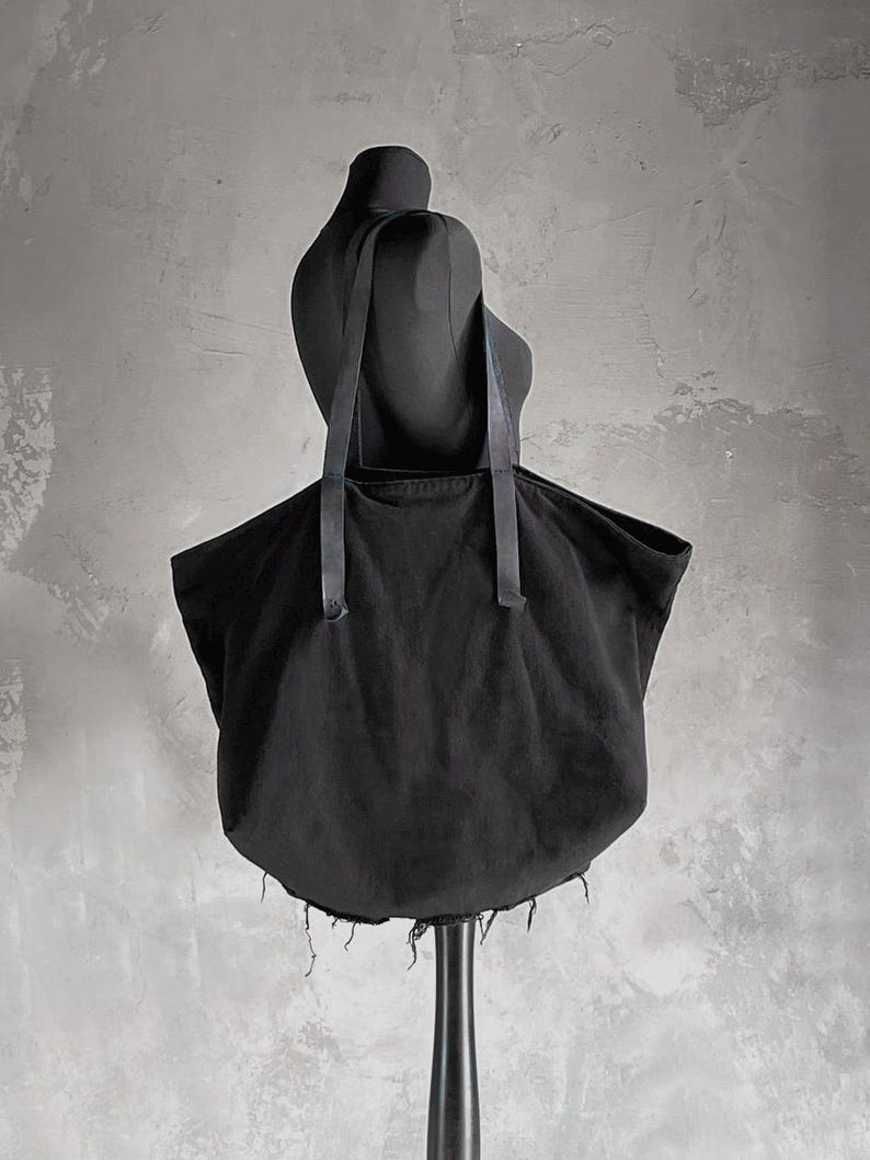 May include: A black tote bag with long straps, displayed on a black mannequin. The bag has a rounded bottom and a frayed edge. The background is a textured gray wall.