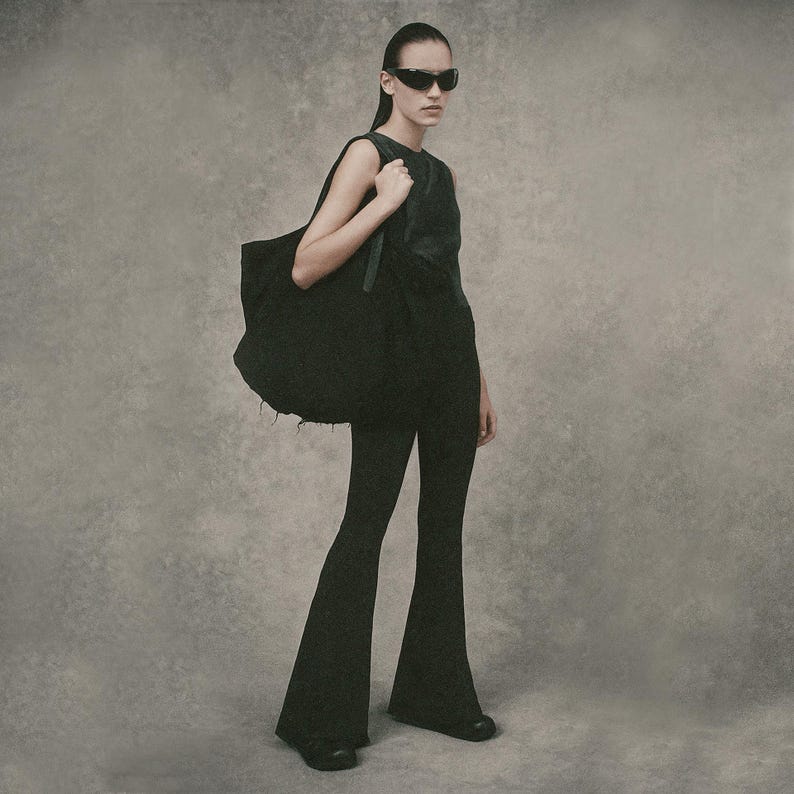 May include: A person wearing black sunglasses, a black sleeveless top, and flared black pants. They are carrying a large black tote bag over their shoulder. The background is a solid, neutral gray.