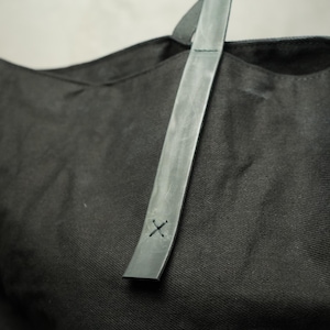 May include: Black tote bag with a gray leather strap. The strap is stitched with a small "X" design. The bag is made of a textured fabric. The strap is attached to the bag.