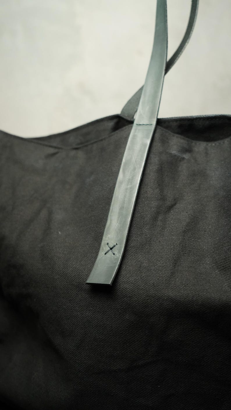 May include: Black tote bag with a gray leather strap. The strap is stitched with a small "X" design. The bag is made of a textured fabric. The strap is attached to the bag.