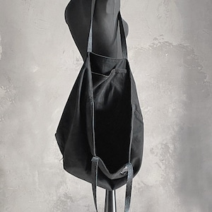 May include: A black tote bag with long straps is displayed on a mannequin. The bag is made of a soft, dark fabric and has a simple, minimalist design. The straps appear to be a contrasting material, possibly leather or a similar texture.