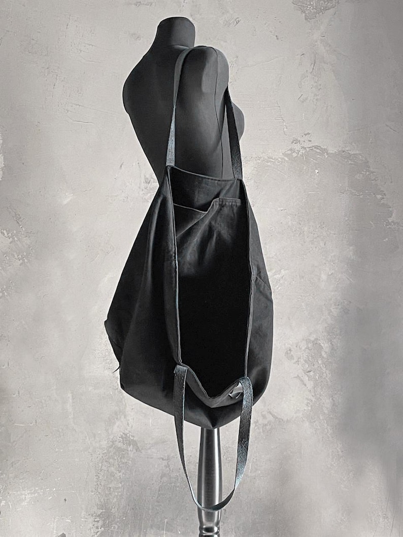 May include: A black tote bag with long straps is displayed on a mannequin. The bag is made of a soft, dark fabric and has a simple, minimalist design. The straps appear to be a contrasting material, possibly leather or a similar texture.
