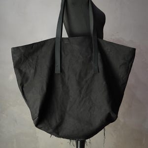 May include: A large black tote bag made of a textured fabric with two long, flat black straps attached to the front. The bag features a wide, open top and a bottom edge with visible frayed, loose threads hanging from the base of the bag.