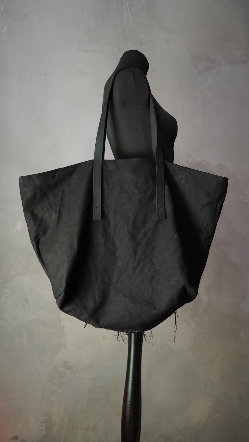 May include: A large black tote bag made of a textured fabric with two long, flat black straps attached to the front. The bag features a wide, open top and a bottom edge with visible frayed, loose threads hanging from the base of the bag.