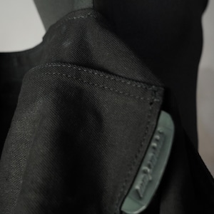 May include: Close-up of a black garment, possibly a jacket or coat, with a visible collar and a small, rectangular label. The fabric appears to be a tightly woven material. The label has text on it.