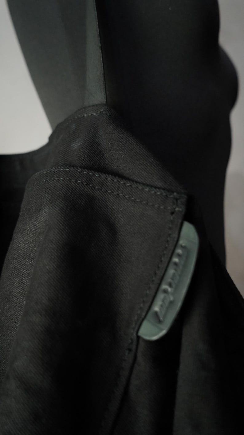 May include: Close-up of a black garment, possibly a jacket or coat, with a visible collar and a small, rectangular label. The fabric appears to be a tightly woven material. The label has text on it.