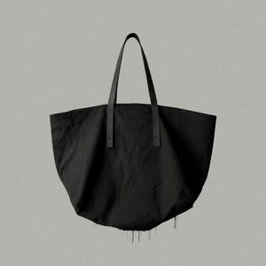 May include: A black tote bag with a wide, rounded body and two long, thin shoulder straps. The straps are attached to the main body with visible cross-stitch detailing. The bottom edge of the bag features a frayed, raw-edge finish.