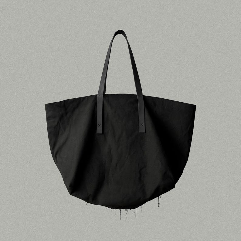 May include: A black tote bag with a wide, rounded body and two long, thin shoulder straps. The straps are attached to the main body with visible cross-stitch detailing. The bottom edge of the bag features a frayed, raw-edge finish.