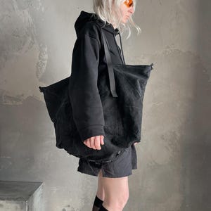May include: A large, unstructured black tote bag made of a textured, matte fabric. The bag features long, thin black shoulder straps and a spacious, slouchy silhouette. The material appears slightly distressed with a deep, solid black colour throughout.