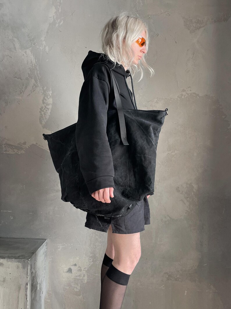 May include: A large, unstructured black tote bag made of a textured, matte fabric. The bag features long, thin black shoulder straps and a spacious, slouchy silhouette. The material appears slightly distressed with a deep, solid black colour throughout.