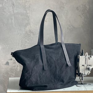 May include: A black canvas tote bag featuring two long, dark leather shoulder straps attached with visible stitching. The bag has a wide, open-top design and a soft, unstructured body, positioned next to a sewing machine on a work surface.