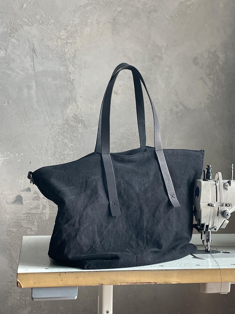 May include: A black canvas tote bag featuring two long, dark leather shoulder straps attached with visible stitching. The bag has a wide, open-top design and a soft, unstructured body, positioned next to a sewing machine on a work surface.