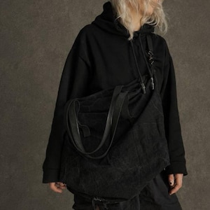 May include: A large black slouchy shoulder bag with a textured surface, featuring two integrated handles. The bag measures 66 cm in width at the top, 47 cm in height, and 20 cm in depth. The bag is worn over a black hooded sweatshirt.