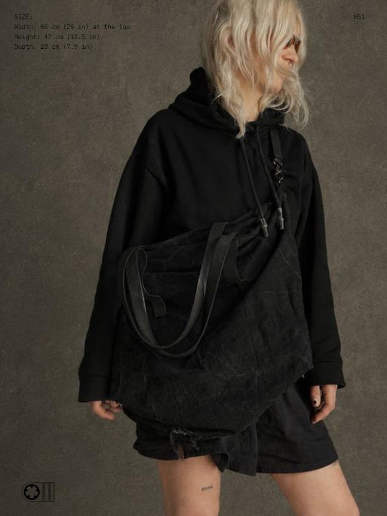 May include: A large black slouchy shoulder bag with a textured surface, featuring two integrated handles. The bag measures 66 cm in width at the top, 47 cm in height, and 20 cm in depth. The bag is worn over a black hooded sweatshirt.